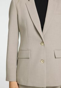 Světlý béžový blazer s upraveným střihem, dvěma předními kapsami a třemi knoflíky. Vyrobeno z hladké texturované tkaniny s jemným vzorem.