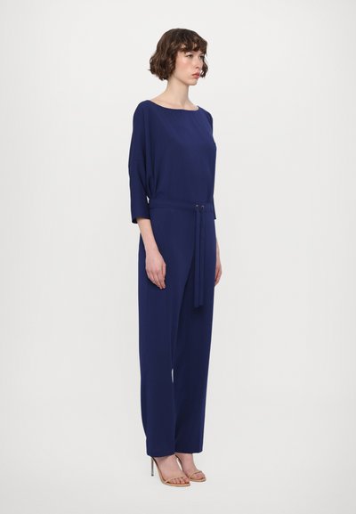 Navy blue jumpsuit with a boat neckline, three-quarter sleeves, and a tie at the waist. Smooth texture, wide legs, and minimal design details.