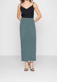 Top en cami noir à volants associé à une jupe maxi en tricot teal ajustée, accentuée par des talons noirs à lanières. Design simple et contemporain.