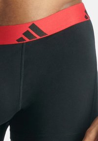 Μαύρα αθλητικά σορτς με κόκκινη ζώνη που διαθέτουν ένα μαύρο λογότυπο. Το ύφασμα είναι λείο με επίπεδες ραφές και εφαρμοστή σχεδίαση.