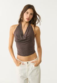 Brauner Trägershirt aus einem strukturierten, glänzenden Material mit einem Cowl-Ausschnitt. Kombiniert mit weißen, hochtaillierten Jeans, die Hände in den Taschen.