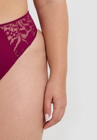 Main droite féminine reposant sur la cuisse, portant de la lingerie en dentelle bordeaux avec des motifs floraux sur un fond clair.
