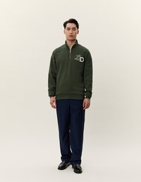 Felpa verde con zip e logo "LD" sul petto, abbinata a pantaloni blu navy. Il modello si trova su uno sfondo neutro, indossando scarpe nere.