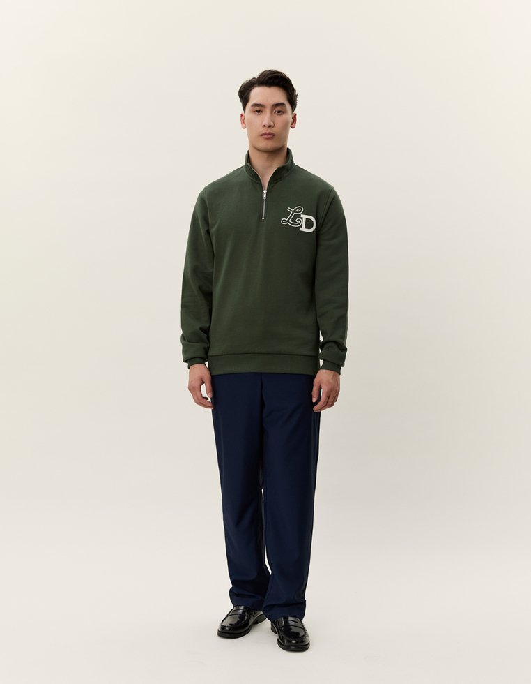 Felpa verde con zip e logo "LD" sul petto, abbinata a pantaloni blu navy. Il modello si trova su uno sfondo neutro, indossando scarpe nere.