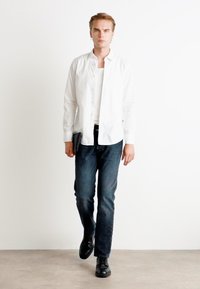 Camisa blanca de botones sobre una camiseta sin mangas blanca, jeans azul oscuro y zapatos negros. El modelo sostiene una tablet oscura y está de pie contra un fondo claro.