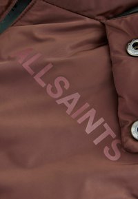 Brunt nylon tyg med "ALLSAINTS" tryckt i en ljusare nyans och en synlig metallknapp som accent. Slät textur, minimalistisk design.