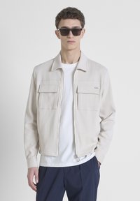 Chaqueta de color beige claro con cremallera y dos bolsillos en el pecho, realizada en material suave, con cuello y acabado texturizado. Llevada sobre una camiseta blanca.
