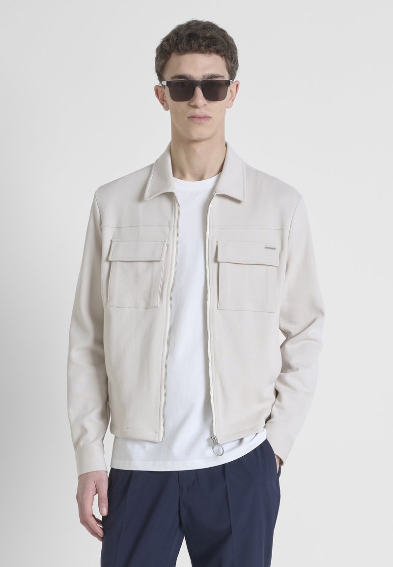 Chaqueta de color beige claro con cremallera y dos bolsillos en el pecho, realizada en material suave, con cuello y acabado texturizado. Llevada sobre una camiseta blanca.