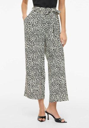 Femme portant un pantalon blanc taille haute à jambes larges avec des pois noirs irréguliers, ceinture nouée à la taille, haut noir et talons ouverts noirs.