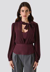 Blusa bordeaux con nodo al collo, maniche lunghe trasparenti, scollatura a V profonda e vita aderente. Abbinata a pantaloni grigi su misura.