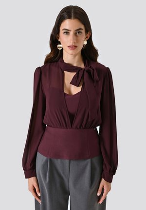 Blusa bordeaux con nodo al collo, maniche lunghe trasparenti, scollatura a V profonda e vita aderente. Abbinata a pantaloni grigi su misura.