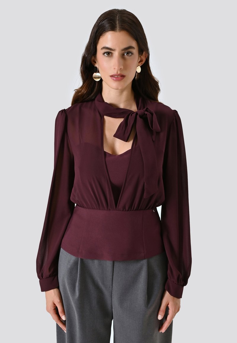 Blusa bordeaux con nodo al collo, maniche lunghe trasparenti, scollatura a V profonda e vita aderente. Abbinata a pantaloni grigi su misura.