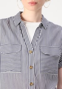Button-up shirt with vertical navy and white stripes, featuring two chest pockets and brown button closures. Collared neckline with short sleeves.