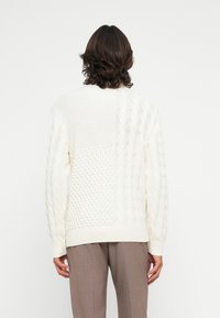 Mann in weißem, strukturierten Strickpullover mit gemischten Mustern, mit dem Rücken zur Kamera, kombiniert mit hellbraunen Hosen vor einem schlichten Hintergrund.