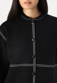 Zwarte gebreide cardigan met een ronde halslijn, opvallende witte stiksels en zwarte knopen aan de voorkant.