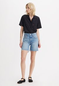 Camisa preta de manga curta com um padrão subtil, combinada com calções de ganga azul claros com bordas desfiadas e cintura alta, sapatos pretos de slip-on.