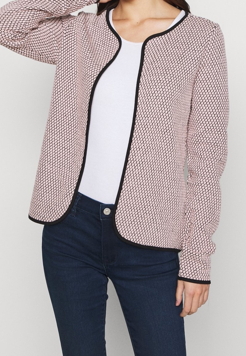 Femme portant un cardigan rose texturé ouvert devant avec bordure noire sur un top blanc et un jean bleu foncé.