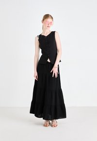 Čierny bezrukávový sveter s prednými gombíkmi, v kombinácii s čiernou maxi sukňou s volánmi. Modelka nosí ružové slnečné okuliare a zlaté sandále. Minimalistický dizajn.