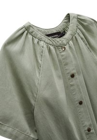 Chemise verte claire à manches courtes, avec un col rond froncé, des boutons sur le devant et une texture côtelée.