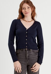 Cárdigan azul marino de manga larga con escote en V y botones dorados al frente. Textura de punto, diseño corto, combinado con unos vaqueros de cintura alta.