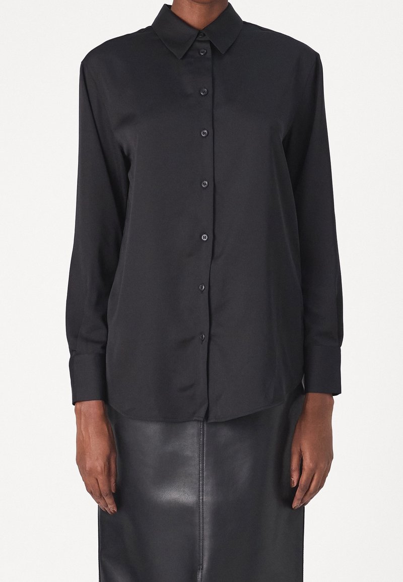Personne portant une chemise noire à manches longues avec col et une jupe en cuir foncé, se tenant devant un fond uni.