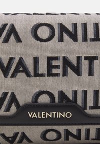 Plátěná kabelka s výrazným černým nápisem "VALENTINO", béžovým pozadím, černými koženými doplňky a zlatými kovovými prvky s logem. Texturovaný látkový povrch.
