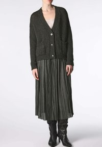Cardigan lavorato a maglia grigio scuro con scollatura a V, chiusura con bottoni e tasche frontali, abbinato a una gonna plissettata verde scuro e stivali neri al ginocchio.