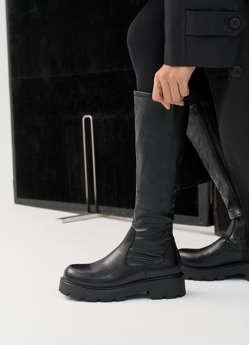 Bottes hautes en cuir noir avec une semelle en caoutchouc épaisse, présentant une finition lisse et un panneau élastique sur le côté pour faciliter l'enfilage.