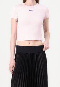 Kvinde iført en lys rosa cropped T-shirt med "HUGO" mærke og højudalte sorte plisserede nederdel, der viser tatoveringer på begge overarme.
