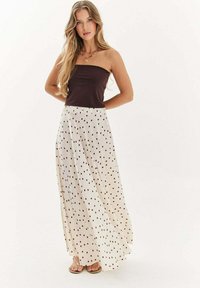 Mladá žena s dlhými blond vlasmi, oblečená v bezramienkovom tmavohnedom topu, bielej sukni s čiernymi bodkami a sandáloch, stojaca na bielom pozadí.