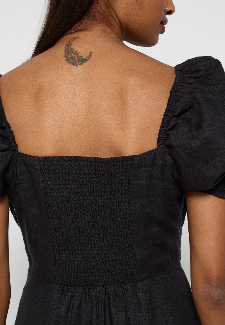 Robe noire avec des manches courtes bouffantes, un décolleté carré et un panneau arrière froncé. Présente une texture lisse et un tatouage floral près de l'épaule.