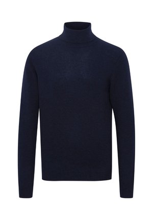 Ung mand iført en marineblå rullekravetrøje, sorte bukser og sorte sko står mod en ensfarvet lys baggrund.
