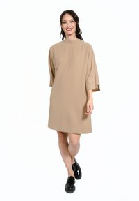 Rochie oversized bej cu un gât înalt și mâneci scurte, realizată dintr-un material neted. Pantofi negri cu talpă platformă.