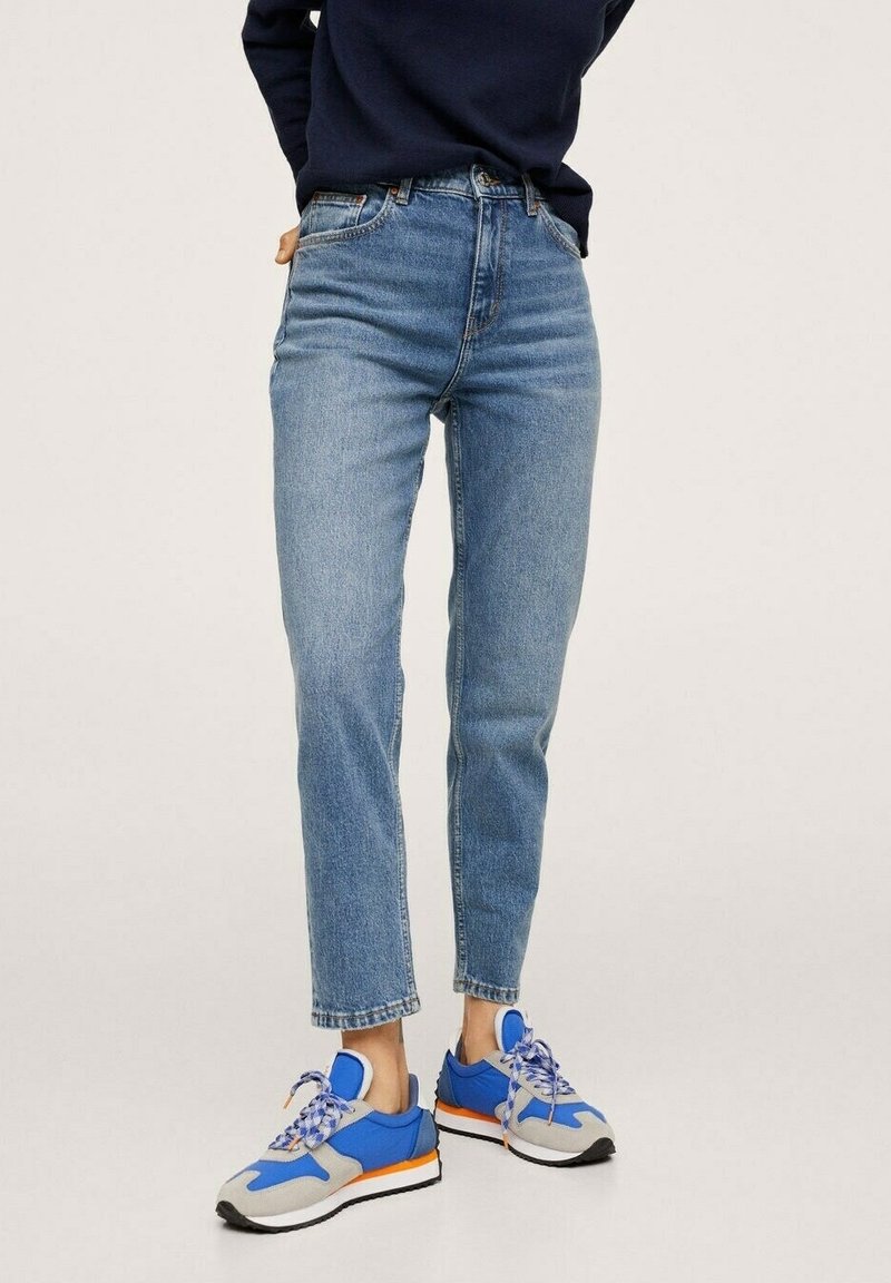 Personne portant un jean taille haute bleu clair, un pull bleu marine foncé, et des baskets bleues, grises, blanches et orange, debout sur un fond uni.