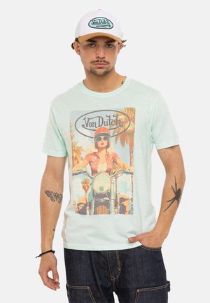 Homme portant une casquette Von Dutch bleu clair et un t-shirt avec un motif vintage de motard, debout avec une main dans la poche.