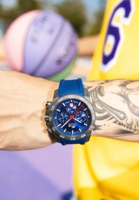 Zils silikona siksnas pulkstenis ar tumšu korpusu, zilu ciparnīcu ar hronogrāfa apakšciparnīcām un sudraba akcentiem. Redzams tetovējums uz plaukstas.