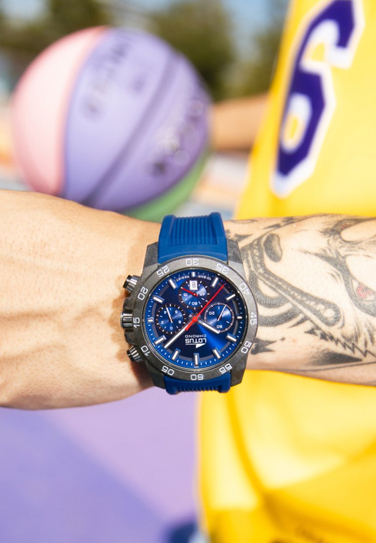 Zils silikona siksnas pulkstenis ar tumšu korpusu, zilu ciparnīcu ar hronogrāfa apakšciparnīcām un sudraba akcentiem. Redzams tetovējums uz plaukstas.