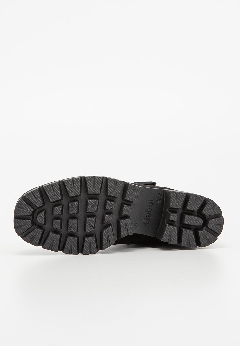 Semelle en caoutchouc noir avec des rainures profondes, présentant une surface texturée et un logo en relief. Talon renforcé et boîte à orteils large pour la stabilité.