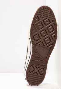 Semelle en caoutchouc marron d'une basket, avec un motif géométrique texturé, comportant une bande blanche sur le côté et un détail de marque indiquant « Converse All Star ».