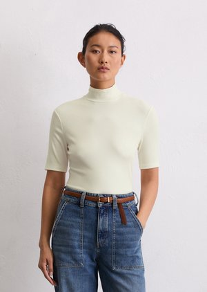 Un haut en crème clair, à col roulé côtelé et manches courtes, associé à un jean en denim bleu à taille haute et coupe décontractée, avec une ceinture en cuir marron.