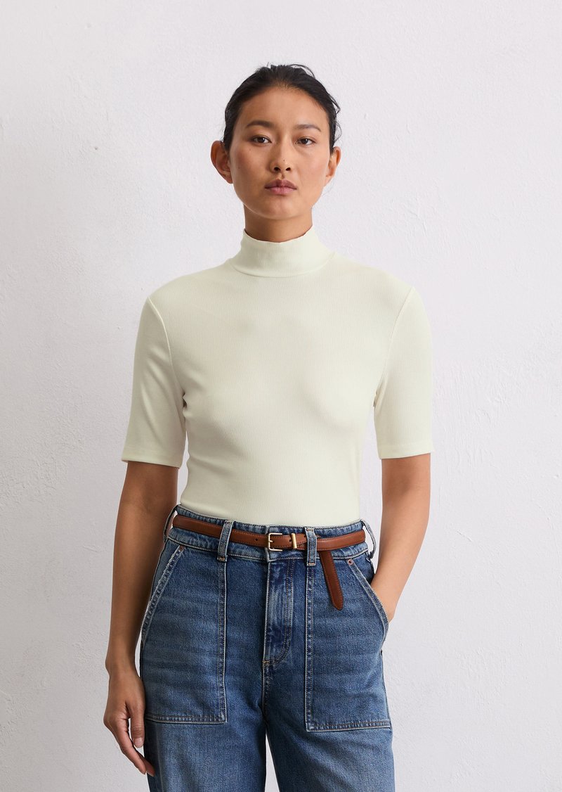 Vaalean kreeminvärinen, ribbiluinen turtleneck-toppi lyhythihaisena, yhdistettynä vyötärön korkeisiin, rentoihin sinisiin farkkuihin ja ruskeaan nahkavyöhön.