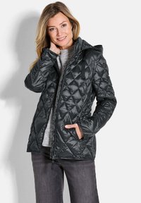 Giacca a vento trapuntata nera con cappuccio, caratterizzata da un motivo diagonale, chiusura con zip e tasche laterali. Indossata sopra un maglione grigio e jeans.