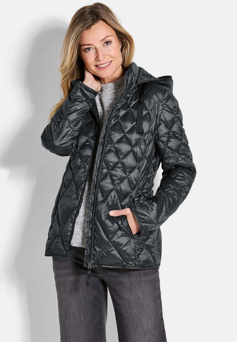 Giacca a vento trapuntata nera con cappuccio, caratterizzata da un motivo diagonale, chiusura con zip e tasche laterali. Indossata sopra un maglione grigio e jeans.
