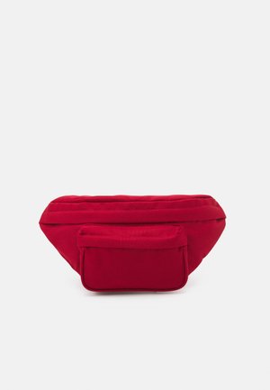 Rote Stoff-Gürteltasche mit einem Hauptfach mit Reißverschluss und einer kleineren vorderen Reißverschlusstasche vor einem einfarbigen Hintergrund.