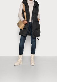 Gilet imbottito nero con cappuccio, borsa a tracolla beige, jeans scuri e stivali in crema con lacci. Texture lisce e elementi di design pratici.