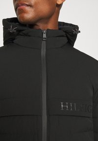 Czarna kurtka puchowa z wysokim kołnierzem, zapięciem na zamek i haftowanym logo "HILFIGER" z przodu. Posiada elegancką, gładką teksturę.