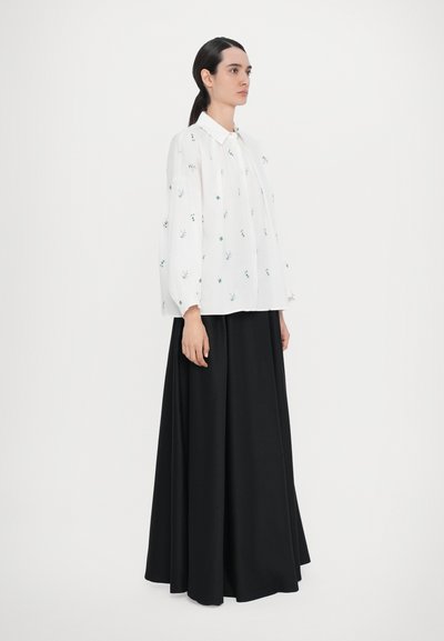 Blouse blanche avec broderie florale verte et coupe ample, associée à une longue jupe noire fluide, créant un contraste de couleurs.