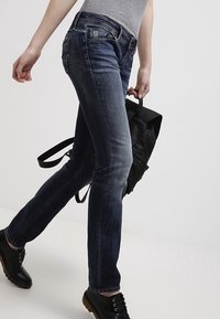Jeans de mezclilla azul oscuro con un diseño de pierna recta, detalles de desvanecimiento y un logo GS Raw en el bolsillo trasero. Combinados con zapatos negros y una mochila.