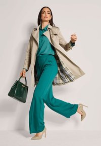 Vrouw loopt zelfverzekerd in een teal pak met een beige trenchcoat, met een donkergroene handtas en beige hoge hakken op een witte achtergrond.