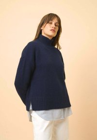 Granatowy sweter z golfem i ściągaczem na dolnej krawędzi oraz bocznymi rozcięciami, nałożony na jasną koszulę w paski. Miękka tekstura, luźny krój.